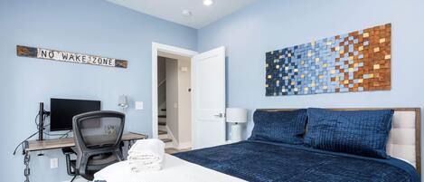 A bedroom with a bed, a desk, a chair, a wall art piece, and a sign that reads 'NO WAKE ZONE'.