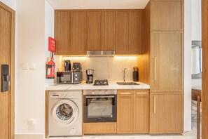 A compact kitchen with wooden cabinets, a built-in washing machine, a coffee maker, and a microwave. - Bloomfields Studio - Azizi Riviera 67-Meydan (Dubai)