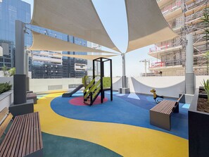Un parque infantil en la azotea con tobogán y estructura de escalada, rodeado de grandes toldos que dan sombra.