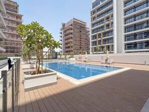 A rooftop swimming pool with a wooden deck, surrounded by modern buildings and some greenery. - Bloomfields Studio - Azizi Riviera 67-Meydan (Dubai)