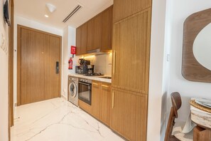 A modern kitchen with wooden cabinets, a built-in oven, a washing machine, and a coffee maker. - Bloomfields Studio - Azizi Riviera 67-Meydan (Dubai)