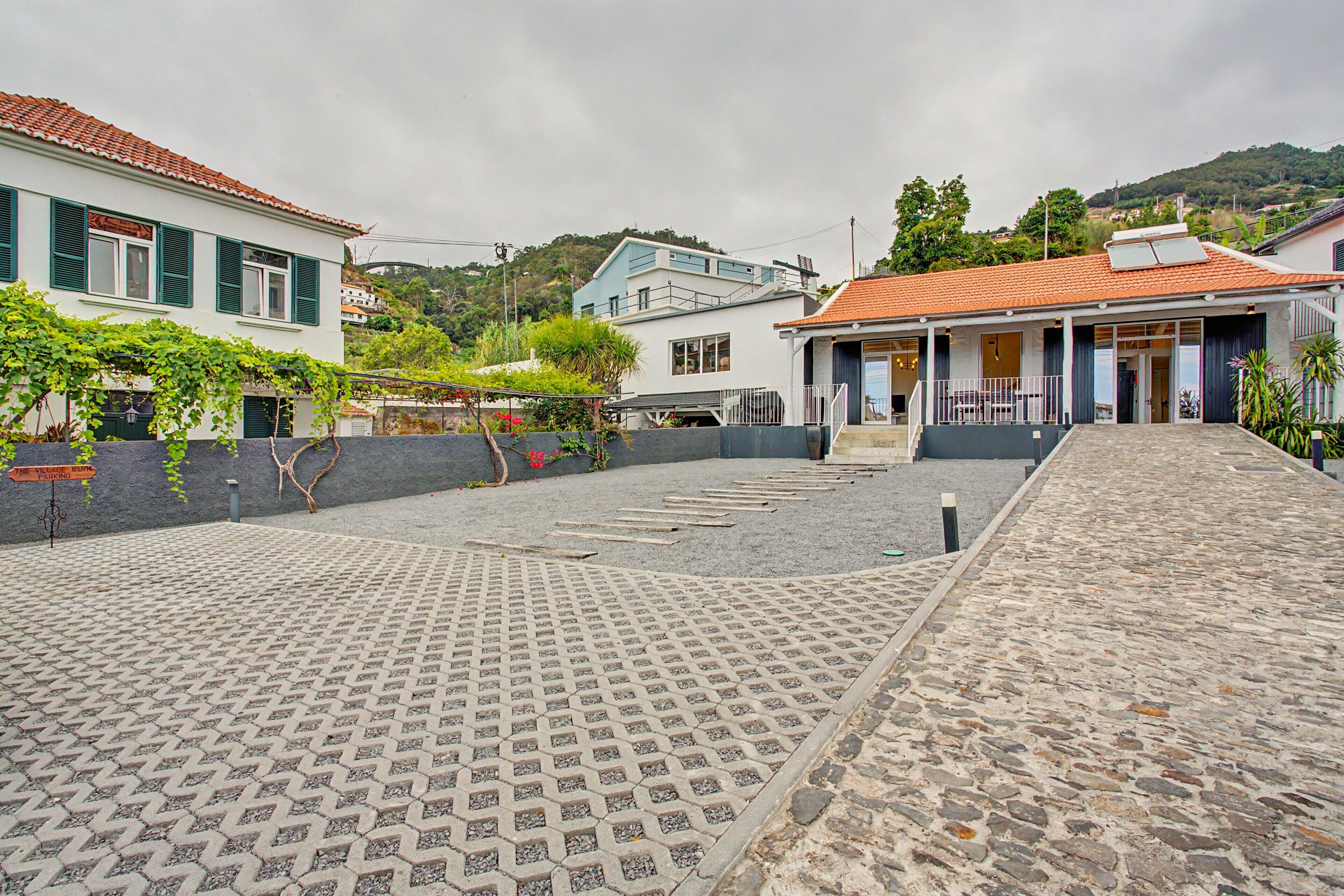 Un patio empedrado con suelo de piedra, un edificio de fachada blanca y contraventanas verdes, y una colina al fondo.
