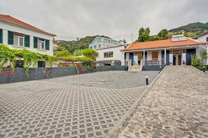 A paved courtyard with a stone floor, a building with a white facade and green shutters, and a hill in the background.