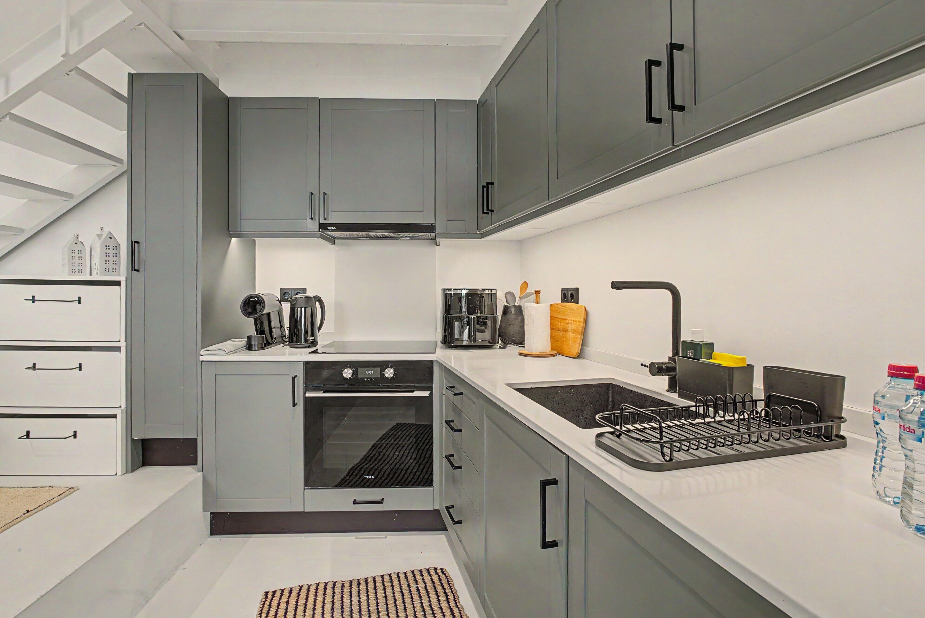 A modern kitchen with grey cabinets, a built-in oven, a sink with a faucet, and a coffee maker.