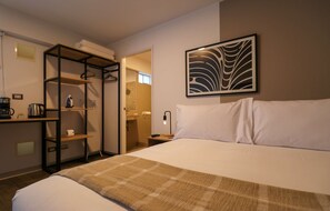 A hotel room with a bed, a desk with a kettle, a shelf, a shower area, and a wall-mounted artwork. - w Cozy Loft in the Heart of Barranco (Lima)