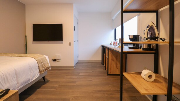 Un dormitorio con cama, televisor montado en la pared, piso de madera y una zona de kitchenette con microondas, hervidor y tabla de planchar.