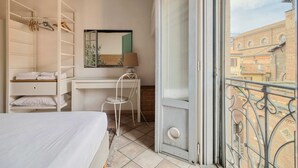 Ein Schlafzimmer mit Bett, Schreibtisch, Stuhl, Kleiderschrank und einem Balkon mit Blick auf Gebäude.
