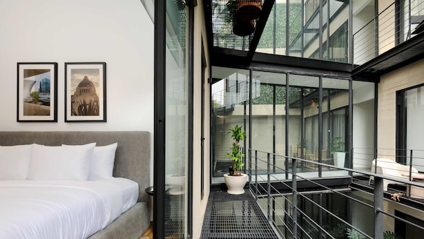 Moderni hotellihuone, jossa on sänky, kaksi kehystettyä kuvaa seinällä, parveke kaiteineen ja ruukussa oleva kasvi.