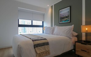Ein Hotelzimmer mit Bett, Nachttisch, Lampe, einem Fenster mit Jalousien und einem gerahmten Bild an der Wand.