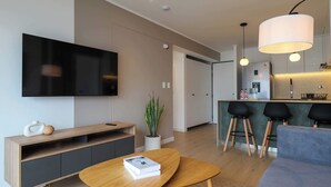 Ein modernes Wohnzimmer mit Flachbildfernseher, einem Holztisch mit einem Buch, einem grauen Sofa, einer Topfpflanze und einer Küchenzeile mit Barhocker und Kühlschrank.