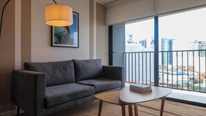 Ein modernes Wohnzimmer mit einem grauen Sofa, einem kleinen runden Tisch, einer Lampe und einem großen Fenster mit Blick auf Gebäude.