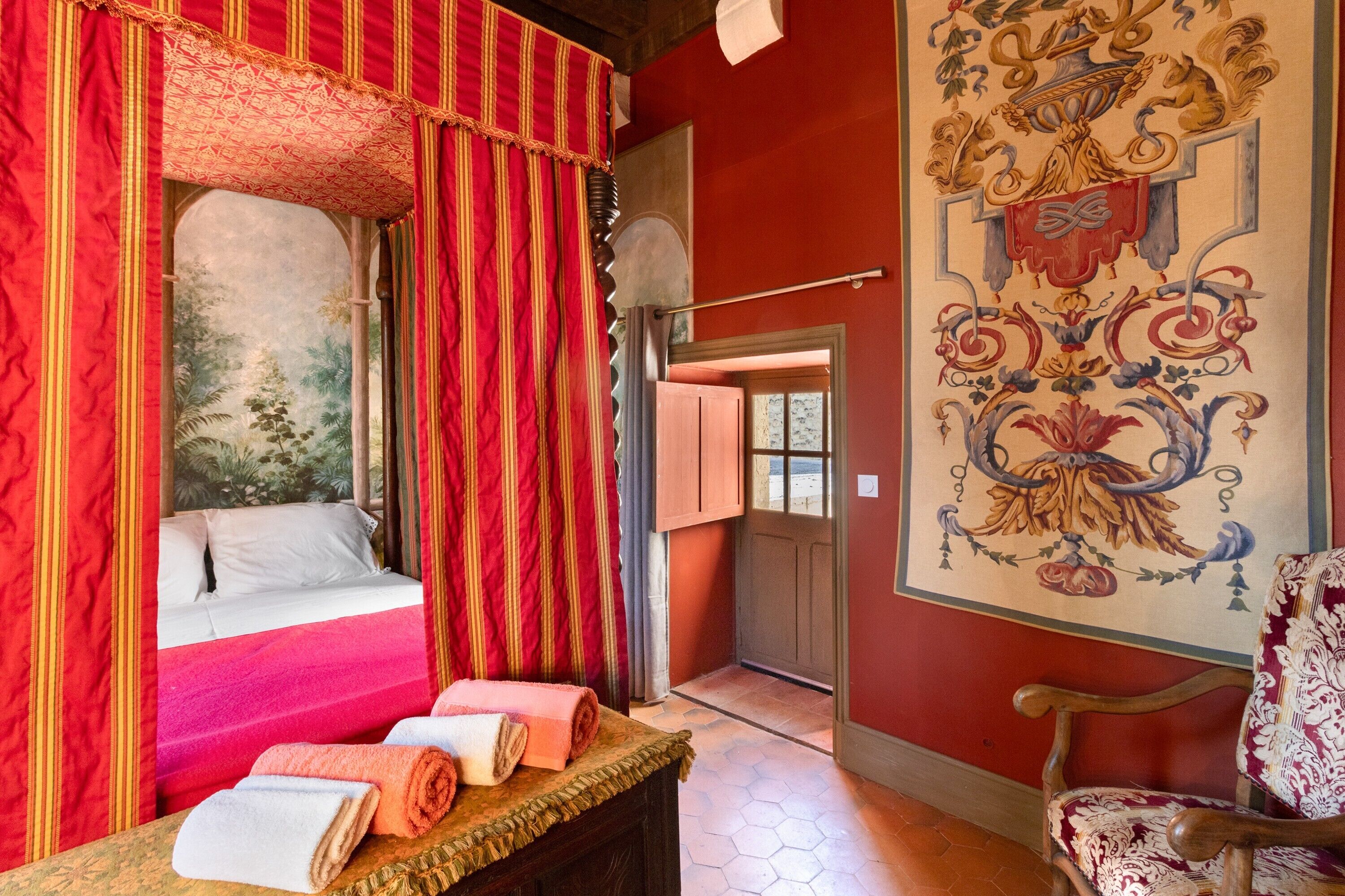 Une pièce avec un lit, des rideaux rouges, une chaise en bois et une tapisserie au mur.