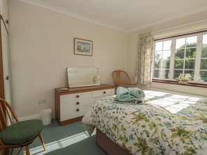 Ein Schlafzimmer mit Bett, Kommode, Stuhl, einem Fenster mit Vorhängen und einem Wandbild.