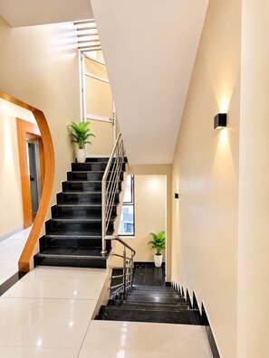 A modern staircase with black steps and a wooden handrail, leading to a landing with a potted plant. - Hotel Grand Dev Krishna (Ujjain)