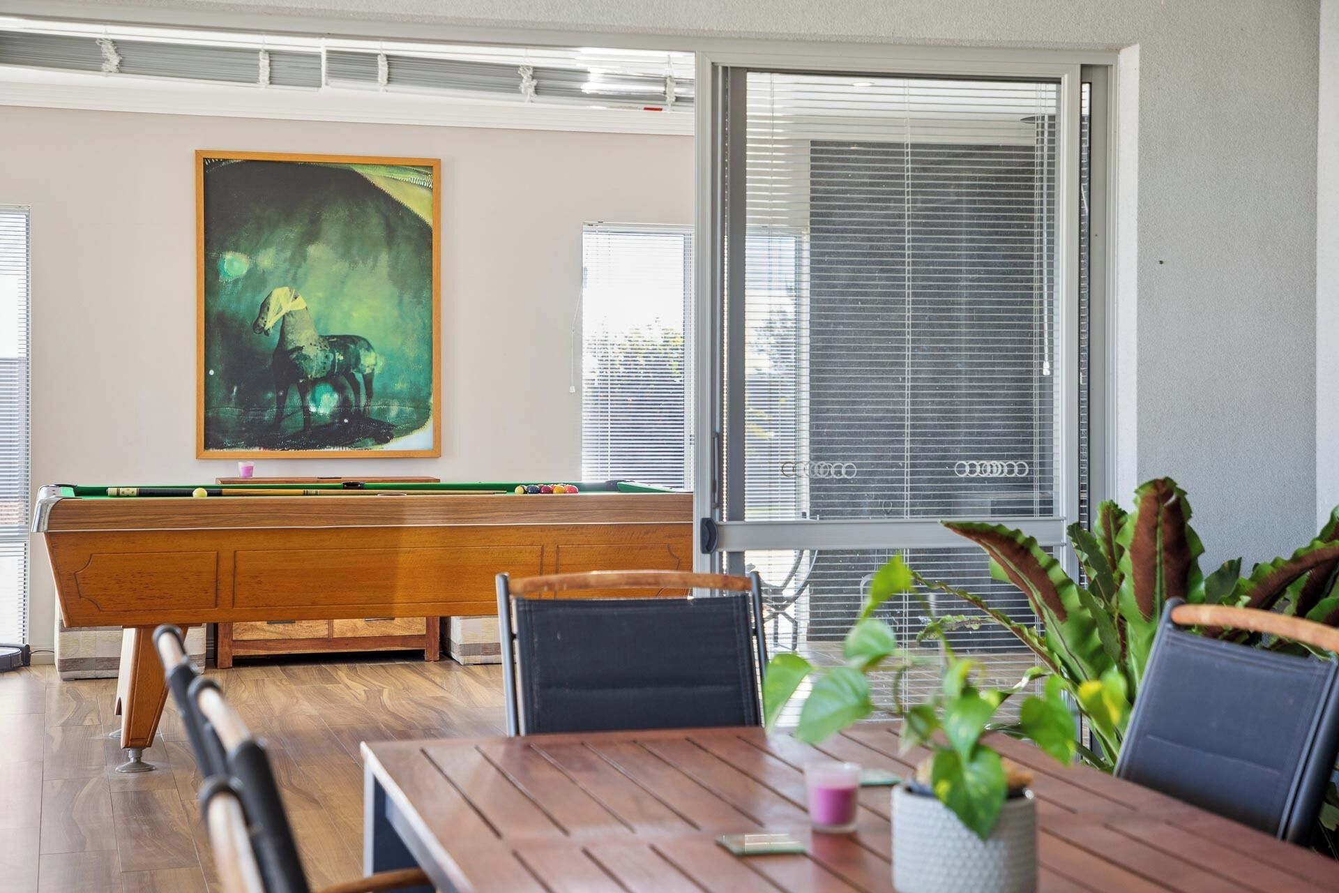 A pool table with a framed abstract painting, a dining table with chairs, and a window with blinds.