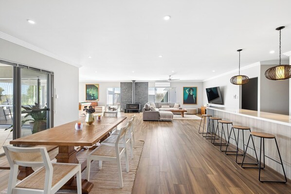 A spacious living area with a wooden dining table, white chairs, a sofa, and a TV.