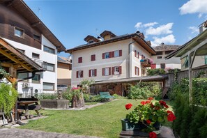 A courtyard with a wooden bench, a stone planter with red flowers, and a building with multiple windows and balconies. - Apartment 'Casa Avisio' with Private Garden, Wi-Fi and Ski Slopes Nearby (Castello-Molina di Fiemme)
