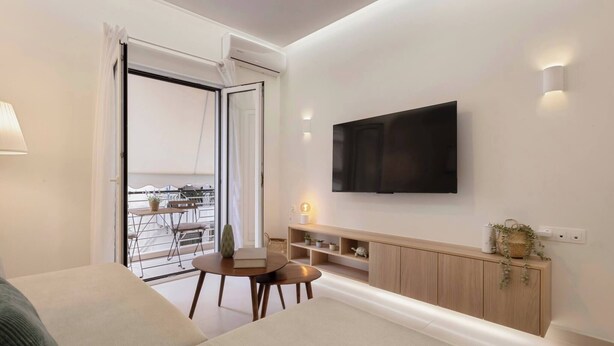 Et moderne værelse med sofa, et lille bord, et fjernsyn monteret på væggen, en balkon med udsigt og et træskab med hylder.
