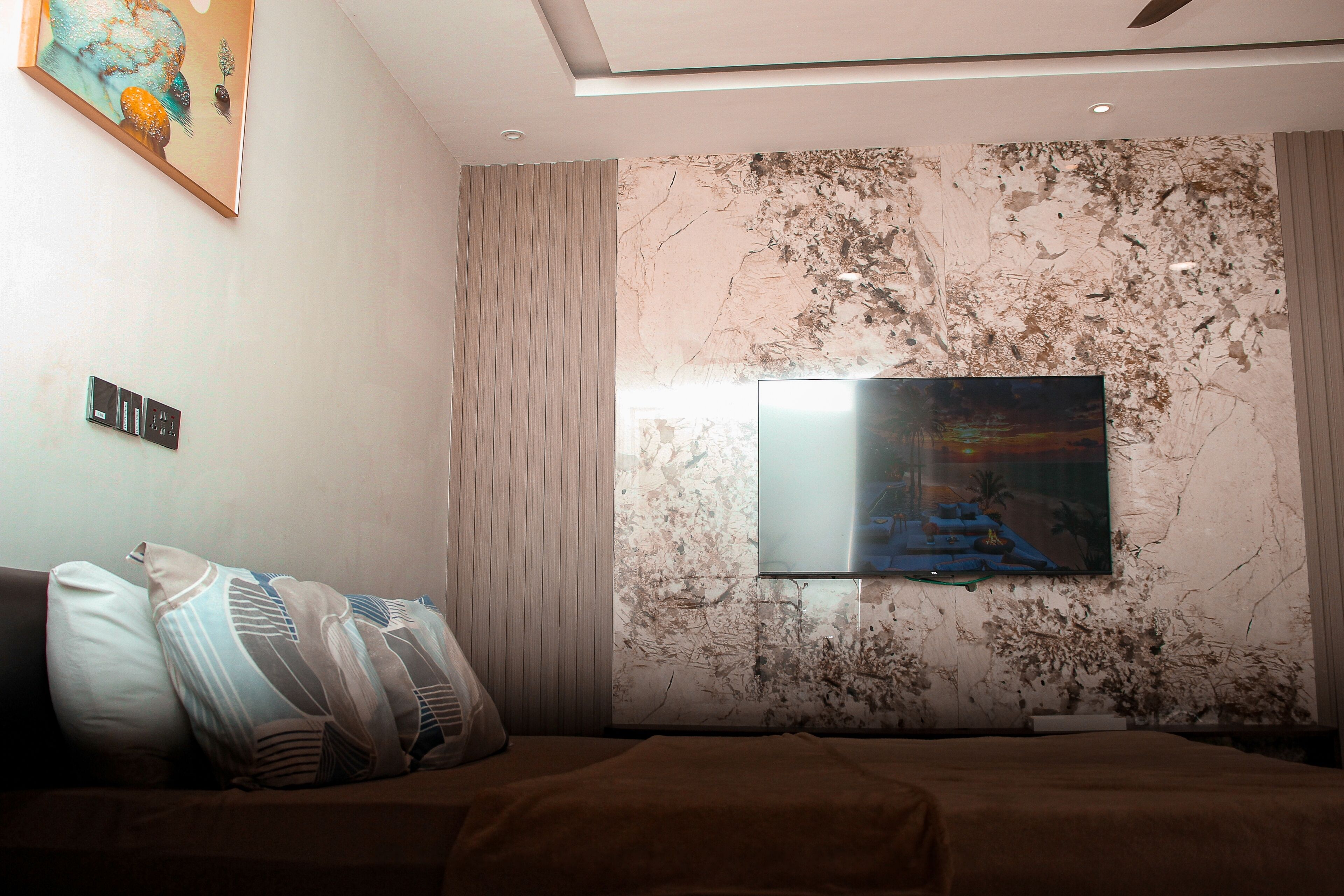 Un dormitorio con cama, almohadas, un televisor montado en una pared texturizada y un cuadro sobre la cama.