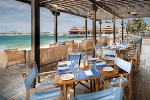 A seaside restaurant with wooden tables and chairs, a view of the ocean, and a building with a red roof in the background. - Luxury 2BR at Anantara Palm Beach & Pool Access (Dubai)