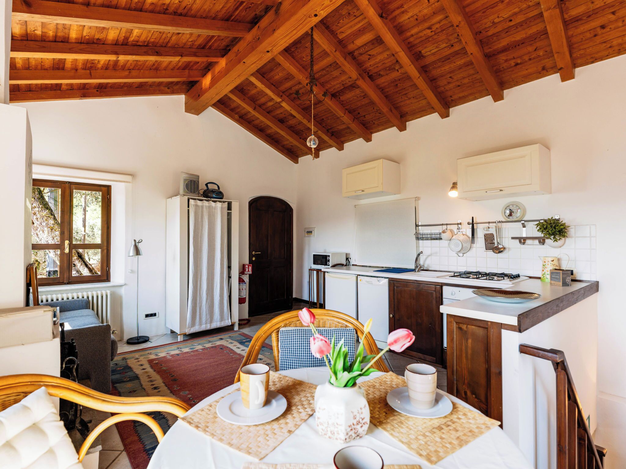 A cozy kitchen with wooden beams, a dining area with a table set for two, and a view of the outside through a window.