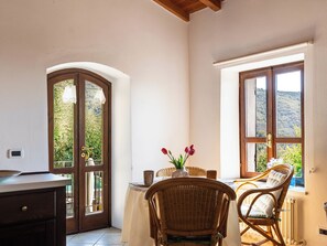 A dining area with a table set for two, a wicker chair, and a view of greenery through a window. - Casetta Pantasina by Interhome (Pantasina)