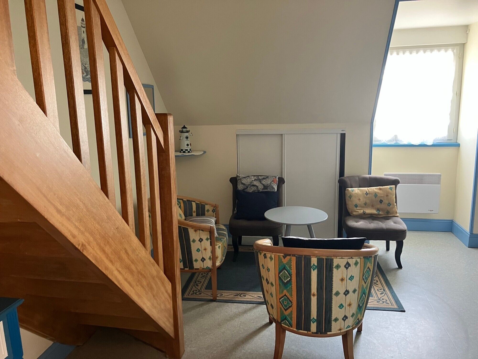 A room with a wooden staircase, a small round table, two chairs with patterned upholstery, a sofa, a decorative wall piece, and a window with blinds.