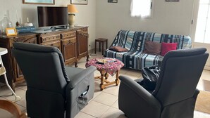Una sala de estar con un sofá, dos sillones, un televisor sobre un mueble de madera, una mesita con una lámpara y cuadros enmarcados en las paredes.