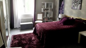 A bedroom with a bed, a chair, a radiator, a shelf with books, and a window with curtains. - Accommodations in St. Bonnet Tronçais (Saint-Bonnet-Tronçais)