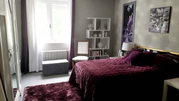 Una camera da letto con un letto, una sedia, un termosifone, uno scaffale pieno di libri e una finestra con tende.