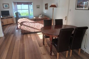 A dining area with a round table and chairs, a sofa, a television, and a window with a view. - The Sailor's Galley (Aghada)