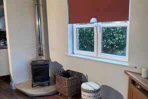 Una habitación con estufa de leña, una ventana con vistas a la vegetación, una cesta de mimbre y un reposapiés estampado.