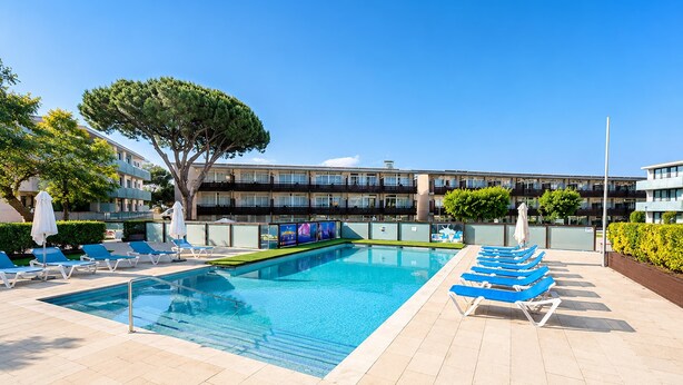 A swimming pool with lounge chairs, a tree, and a building with balconies in the background. - Romantic Coastal Apartment Hideaway (Castell-Platja d'Aro)