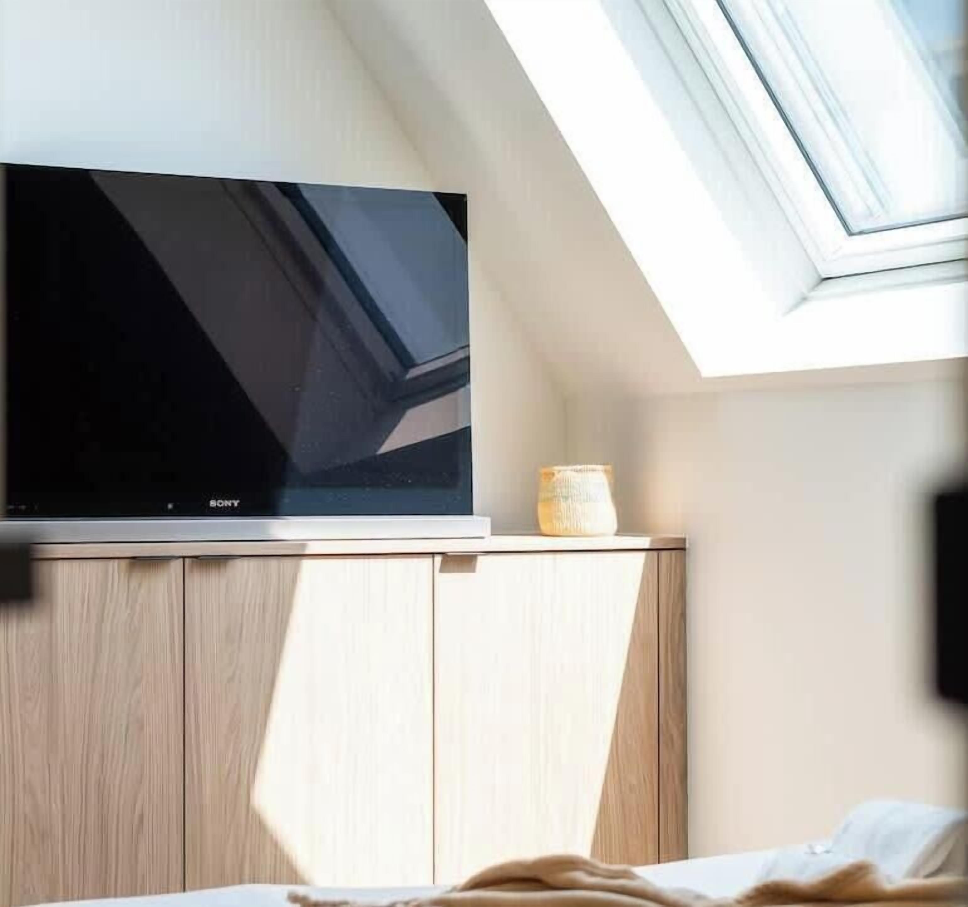 A modern room with a flat-screen TV mounted on a wooden cabinet, a small decorative vase, and a bed with a white blanket.