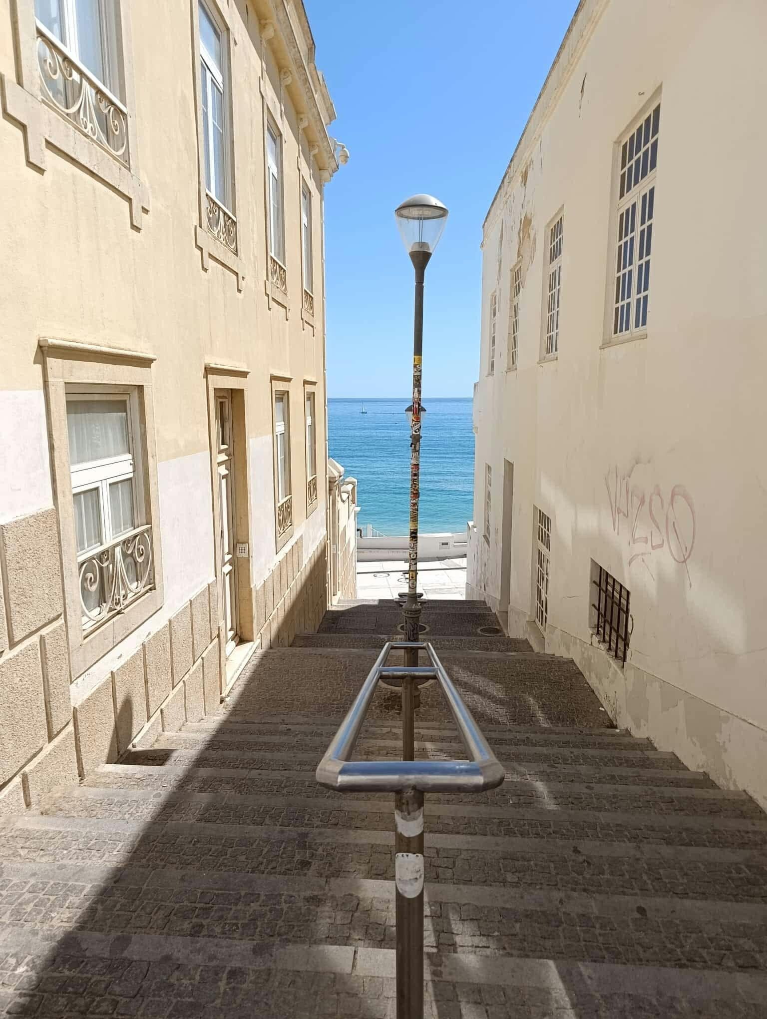 Une rue étroite, bordée d’une rambarde en métal et d’un lampadaire, qui mène à un immeuble offrant une vue sur la mer.