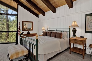 Een slaapkamer met een bed, twee nachtkastjes, een stoel, een raam met uitzicht op bomen en een ingelijste botanische illustratie aan de muur.
