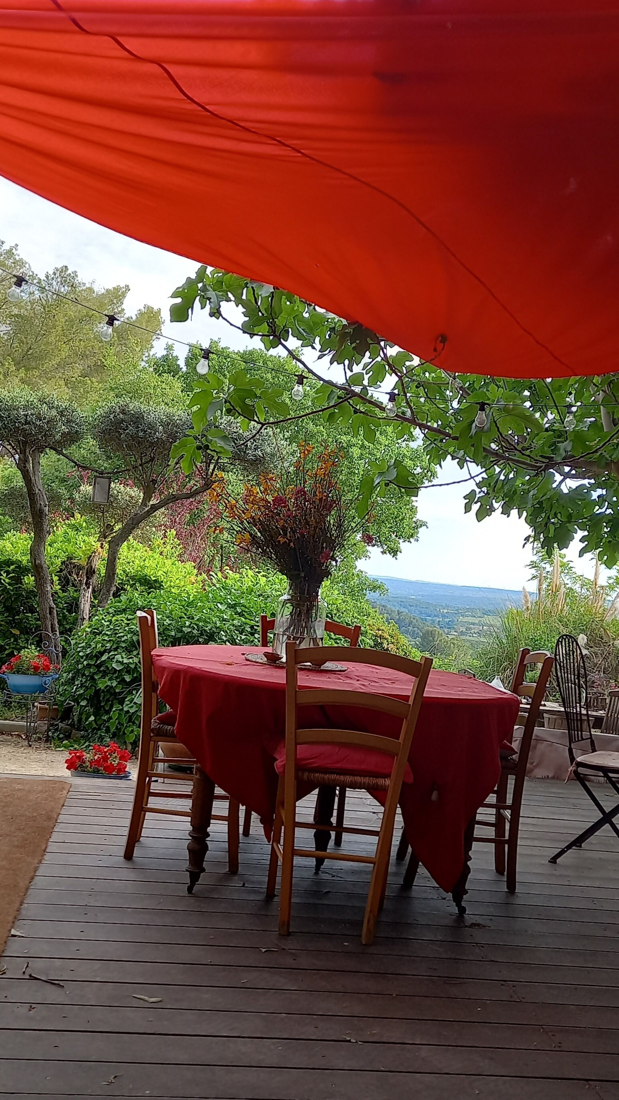 An outdoor dining area with a red table and chairs, surrounded by greenery and a view of the landscape.