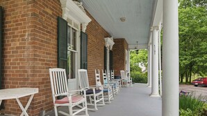 A covered porch with white rocking chairs and a table, brick walls, and columns.