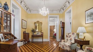A room with a wooden parquet floor, a chandelier, a floral armchair, a wooden cabinet, and a mirror.