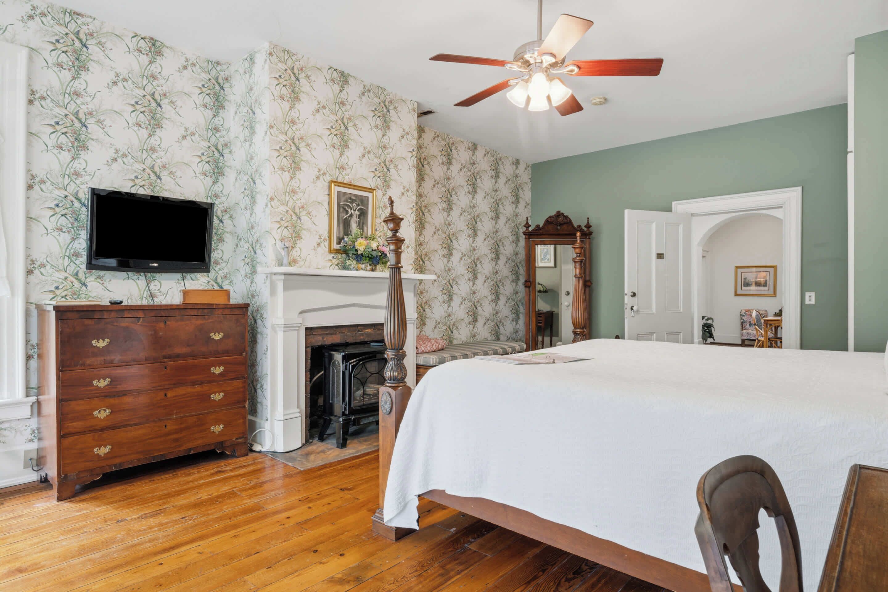 A bedroom with a bed, wooden dresser, television, fireplace, and a ceiling fan.