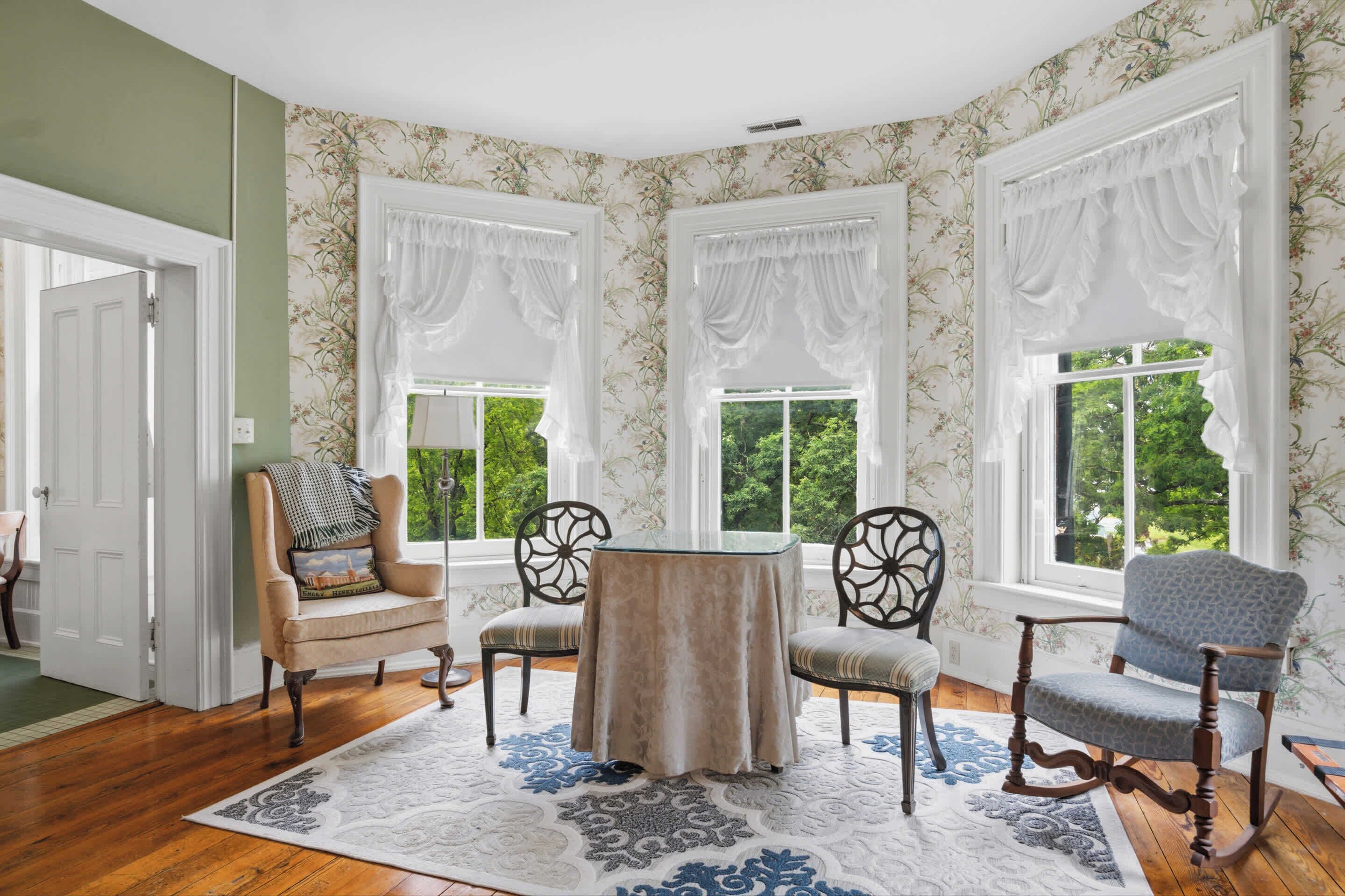 A room with a round table, two chairs, a rocking chair, and three windows with white curtains.