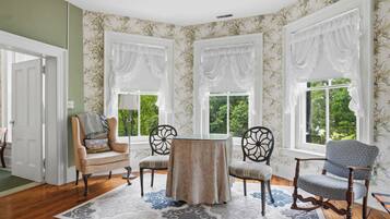 A room with a round table, two chairs, a rocking chair, and three windows with white curtains.