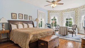 A bedroom with a bed, bedside tables, a wooden chest, a ceiling fan, and a window with curtains.