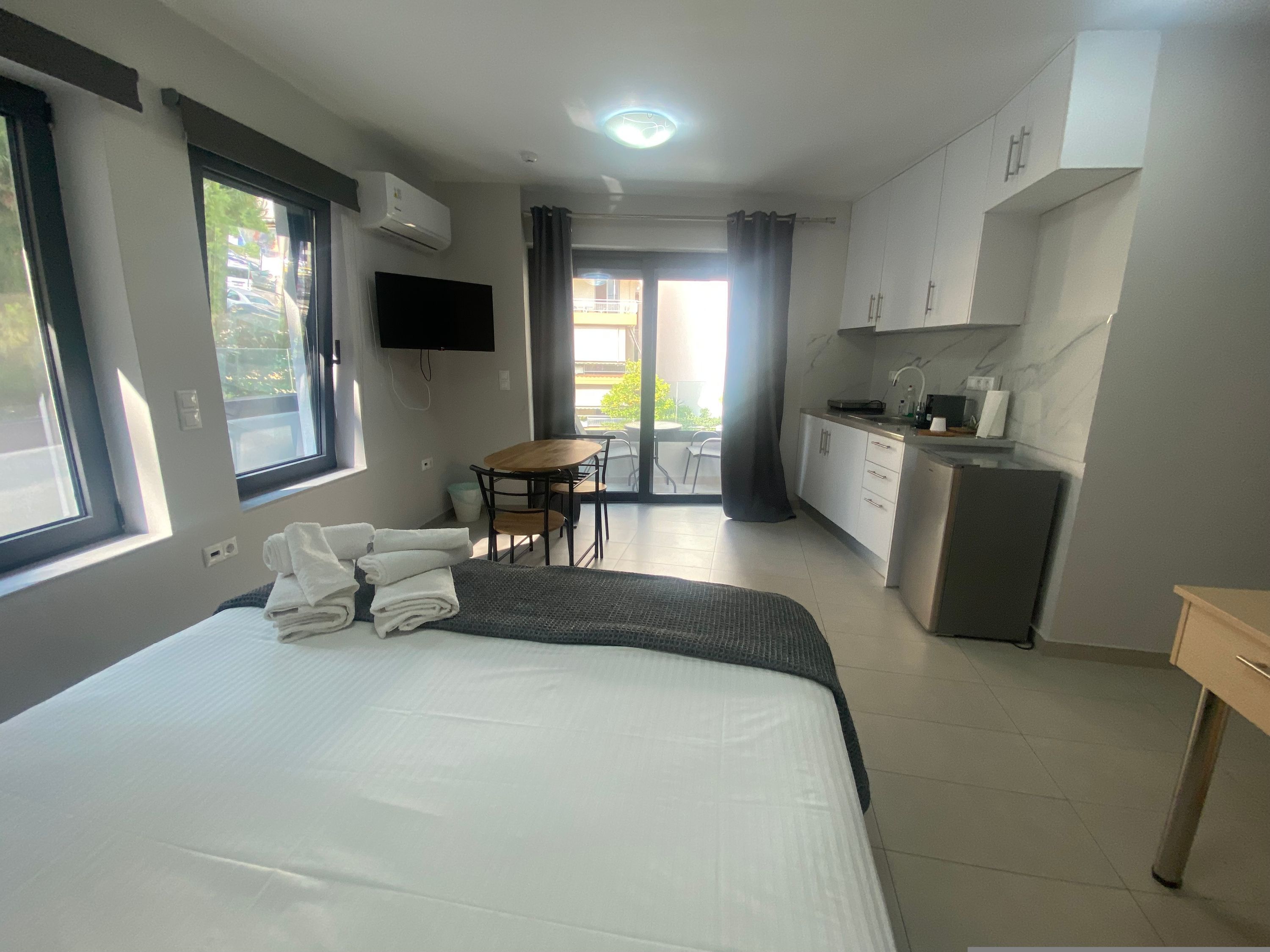 Une chambre d’hôtel équipée d’un lit, d’un bureau, d’une chaise, d’une télévision, d’une kitchenette et d’un balcon avec des rideaux.