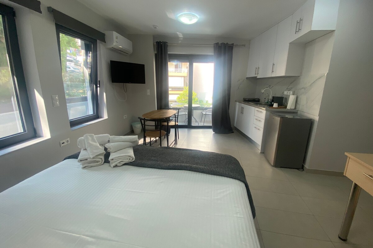 A hotel room with a bed, a desk, a chair, a TV, a kitchenette, and a balcony with curtains.