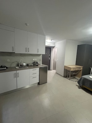 Cozinha compacta com armários brancos, uma geladeira pequena e um espaço de refeições com mesa e cadeira.