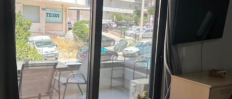 A room with a wooden table, a potted plant, a TV, and a view of a street with parked cars and a building with a sign.