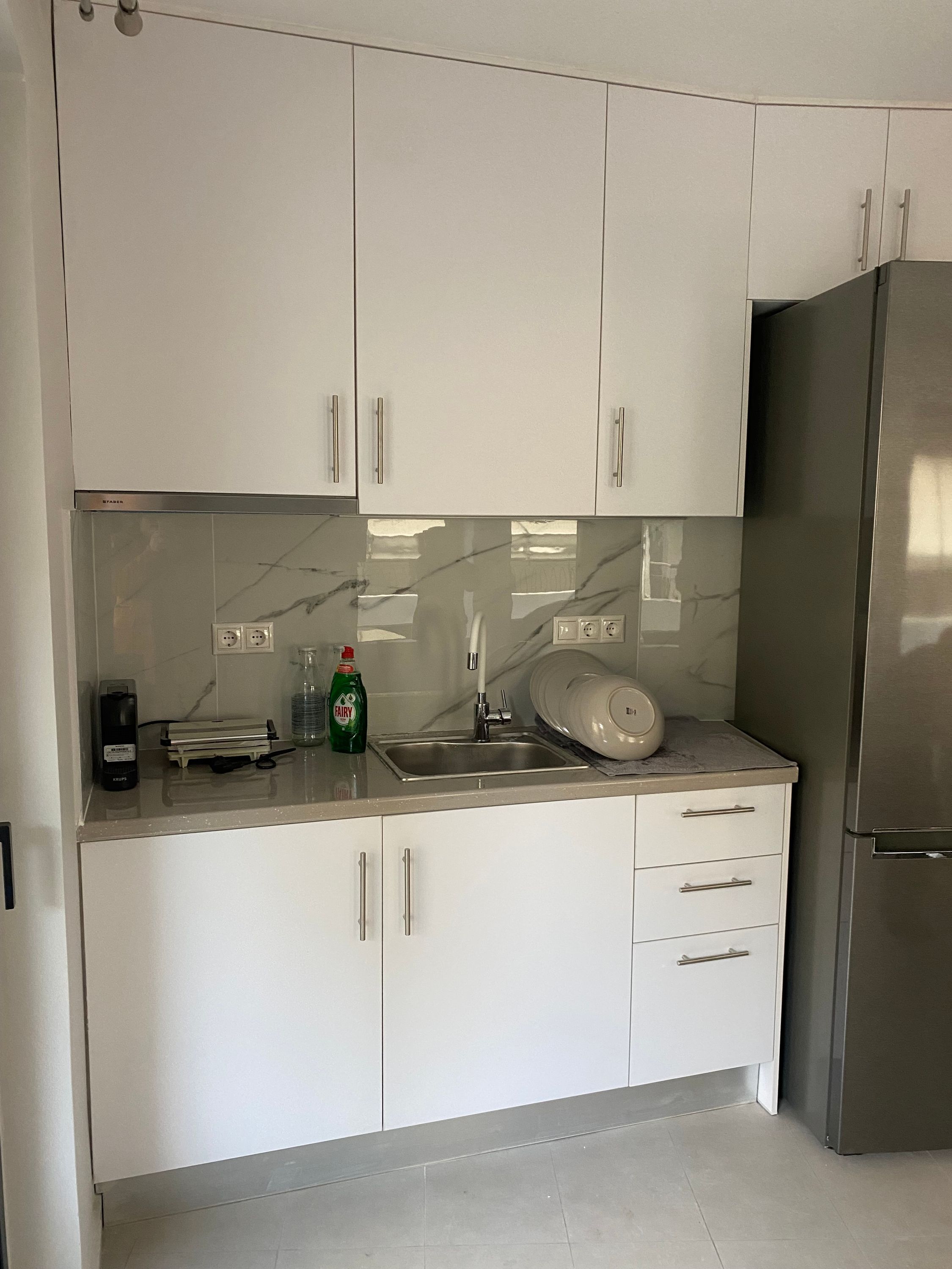 A modern kitchen with white cabinets, a stainless steel refrigerator, a sink, and a countertop with a dish soap bottle.