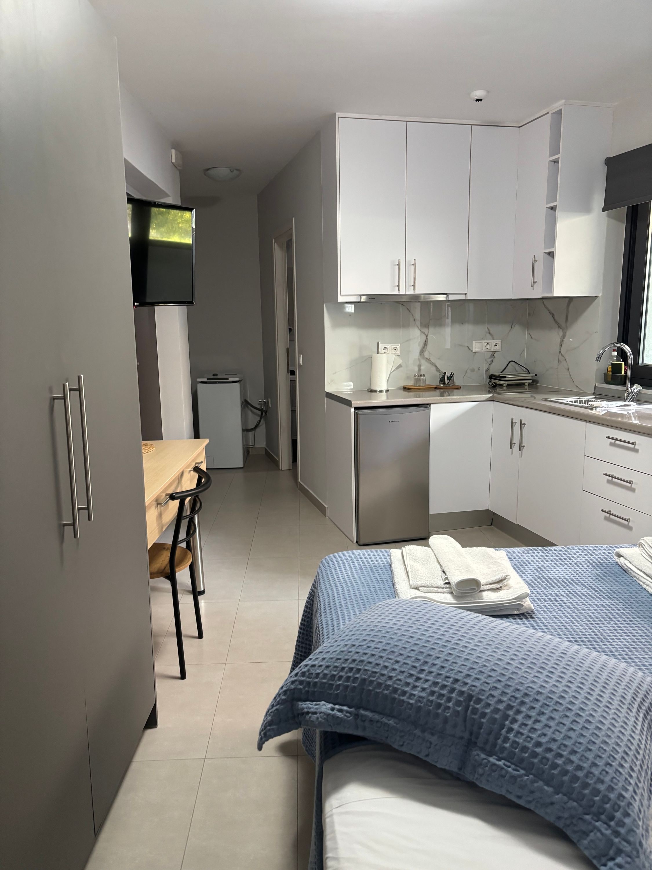 A modern hotel room with a bed, a desk, a kitchenette, and a TV mounted on the wall.