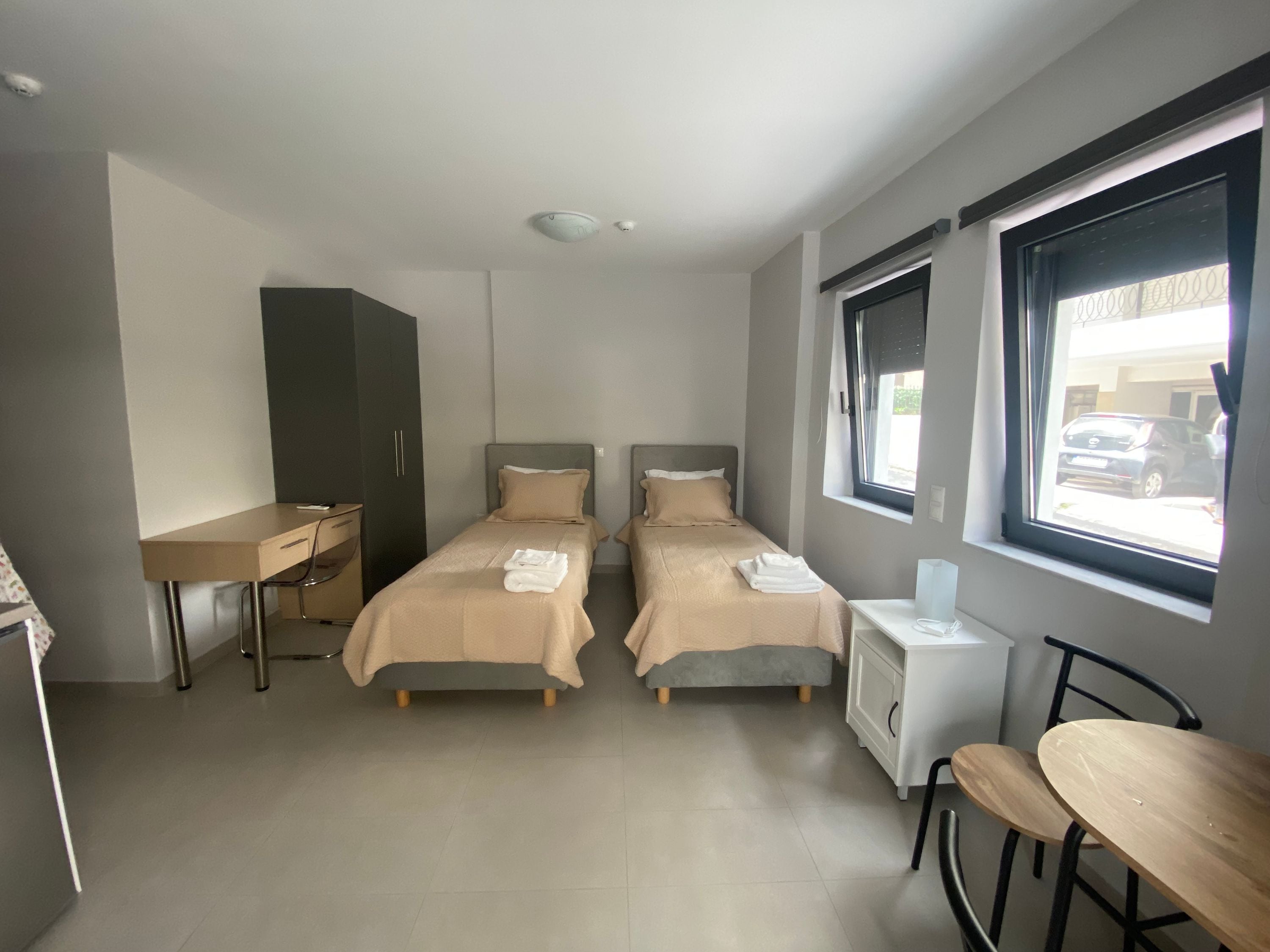 A twin bedroom with two beds, a desk, a chair, and a window with a view of the street.